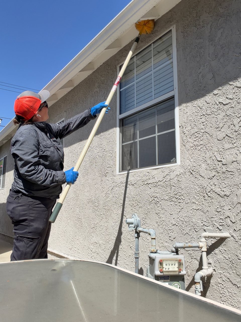 Chris using an extension brush to clear spider webs from the eaves of a stucco house in La Puente